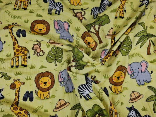 A light yellow-green double gauze fabric with a colorful safari animal print, including cartoon lions, giraffes, zebras, elephants, monkeys, palm trees, and hats.
